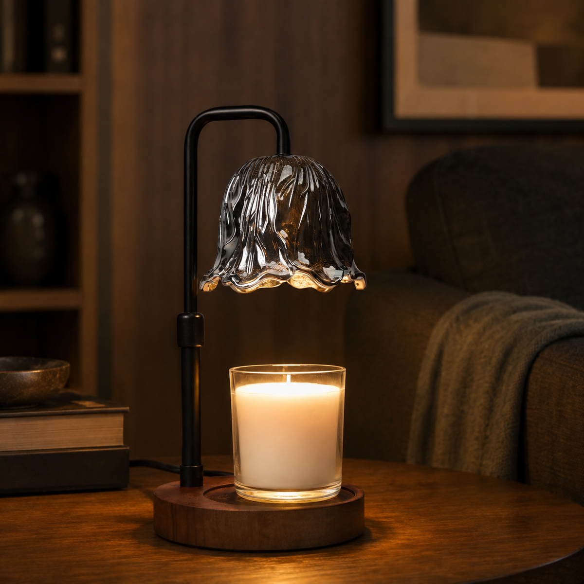 Illuminated candle warmer with dark floral glass shade on a wooden side table beside books and a dark armchair.