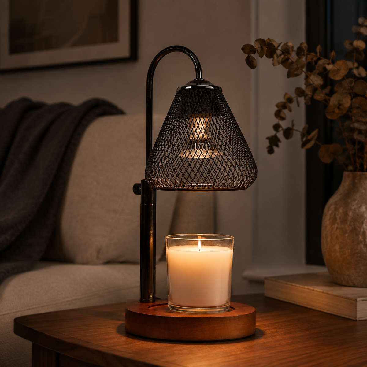 Black mesh candle warmer illuminated on a dark wooden side table beside a sofa and dried foliage.
