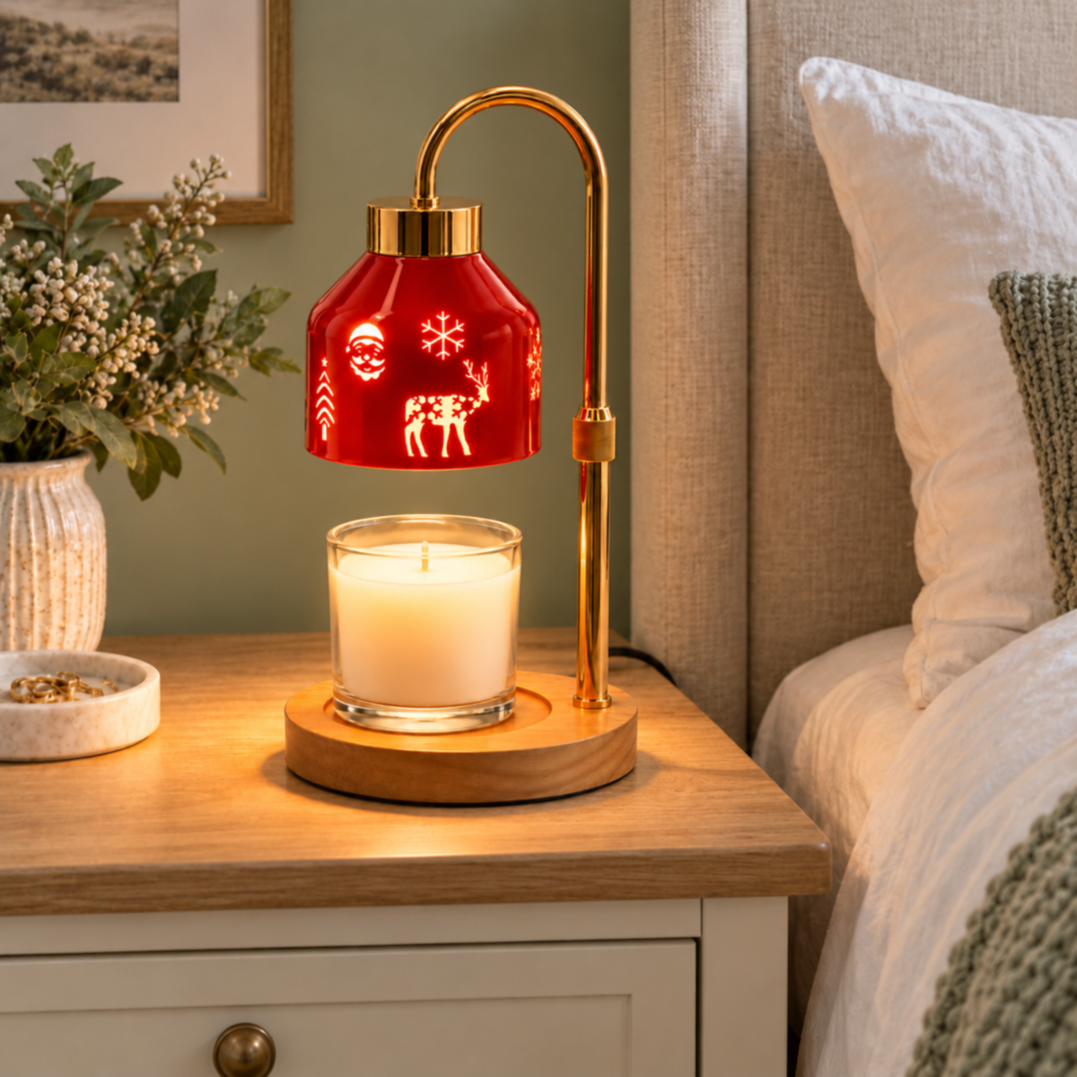 Illuminated red Christmas candle warmer lamp with a clear glass jar candle on a wooden bedside table beside an upholstered bed.
