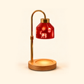 Illuminated red Christmas candle warmer lamp with a curved gold stand and an empty round wooden base on a cream background.