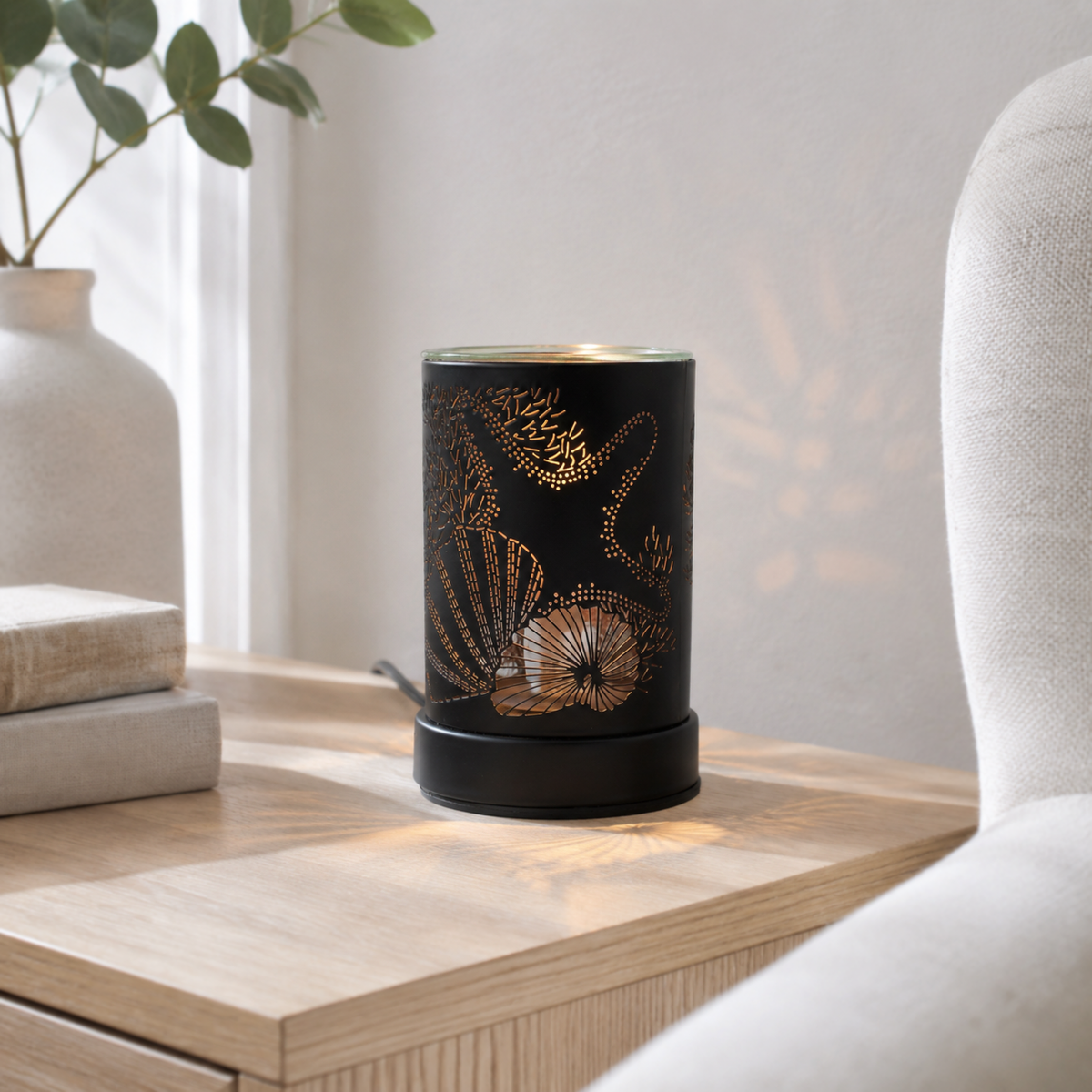 Coastal tabletop wax warmer illuminated on a light wood side table beside books and a white armchair.
