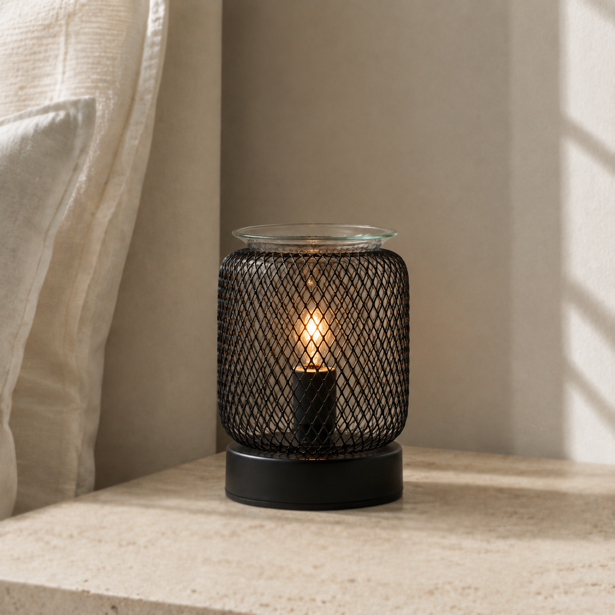 Tall mesh tabletop wax warmer glowing softly on a pale stone ledge beside a sheer curtain.
