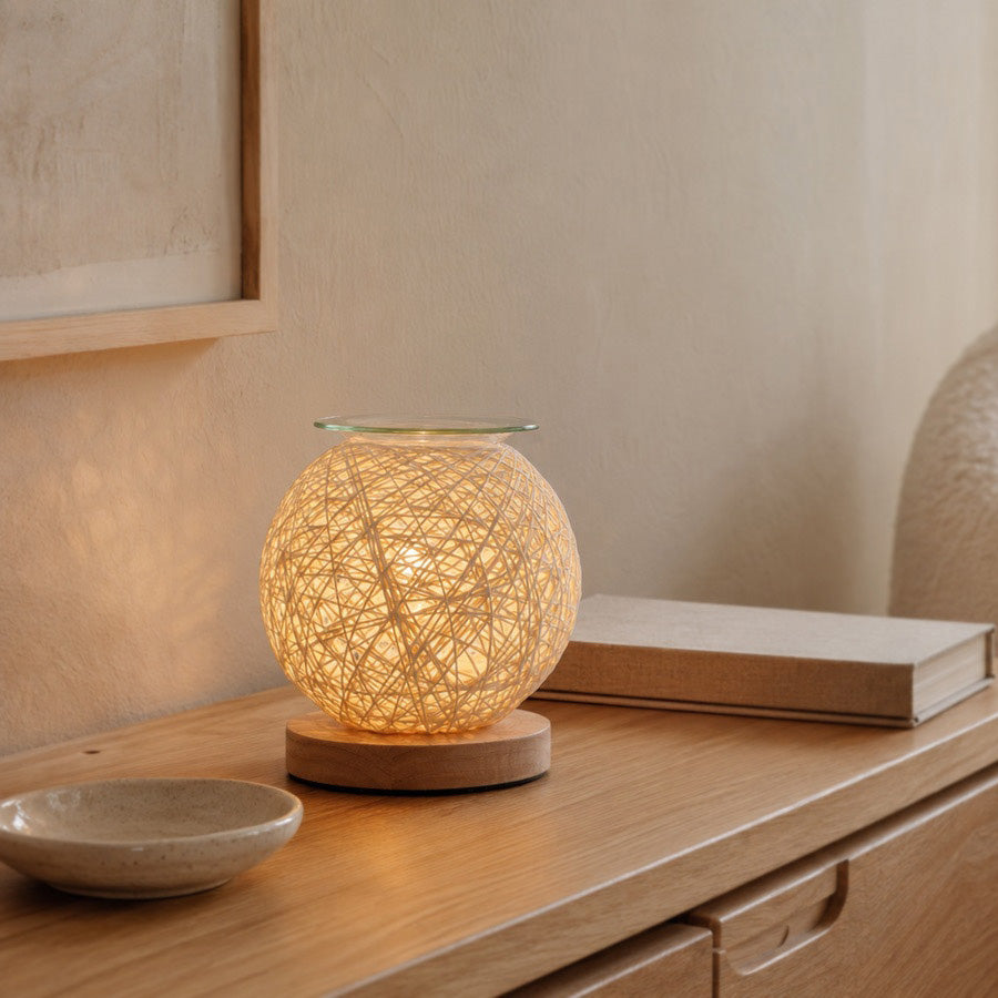 Woven tabletop wax warmer glowing on a wooden console beside a ceramic dish and closed book.