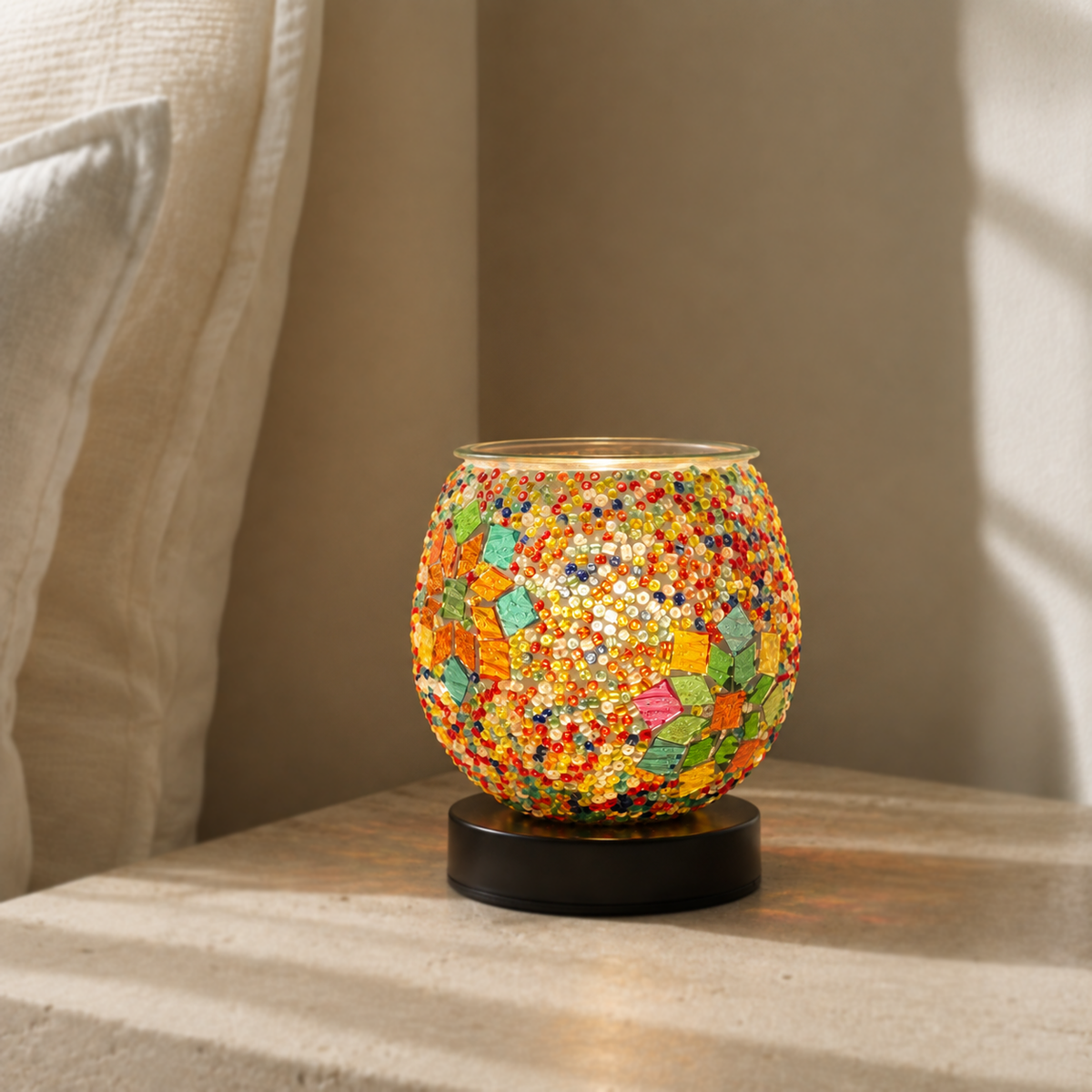 Mosaic glass electric wax warmer softly illuminated on a pale stone surface beside a sheer curtain.