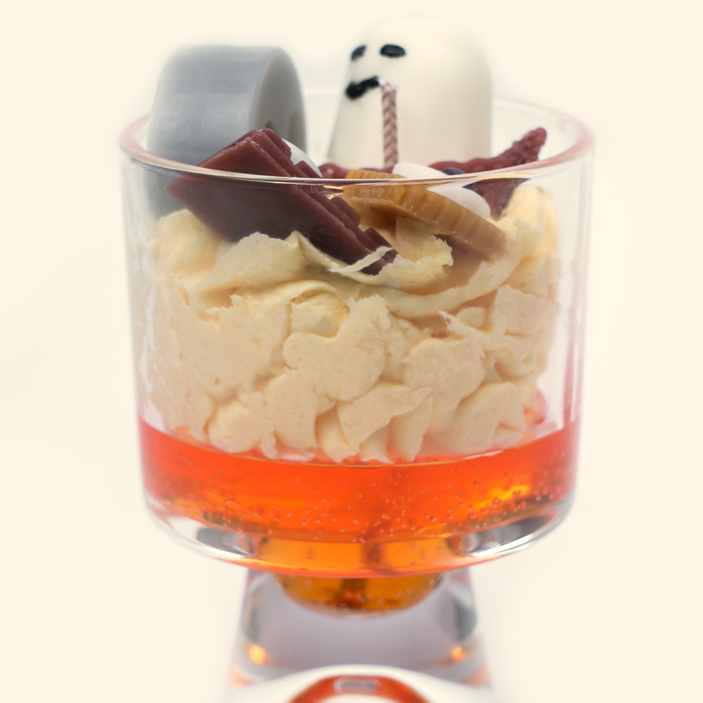 Side view of the footed glass candle with gray and white ghost figures above cream piped wax, brown bar and biscuit-shaped details, and an orange-red gel layer.