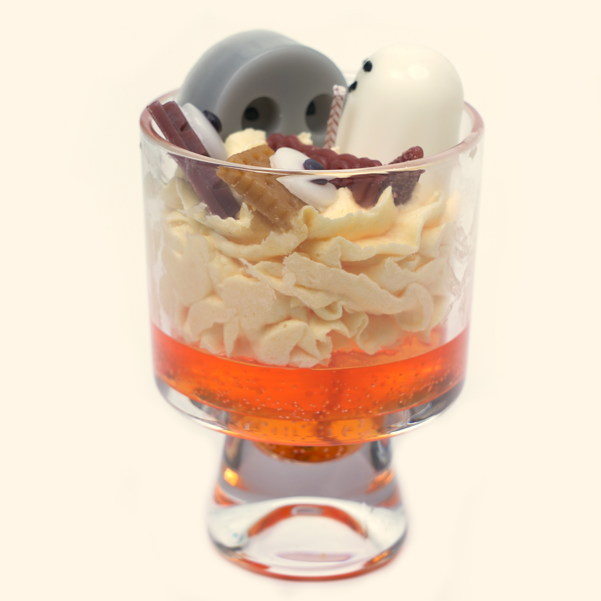 Little Ghost’s Party Halloween Snow Candle in a tall footed glass cup with gray and white ghosts, eyeball-shaped wax pieces, brown candy details and cream piped wax over an orange-red gel layer.