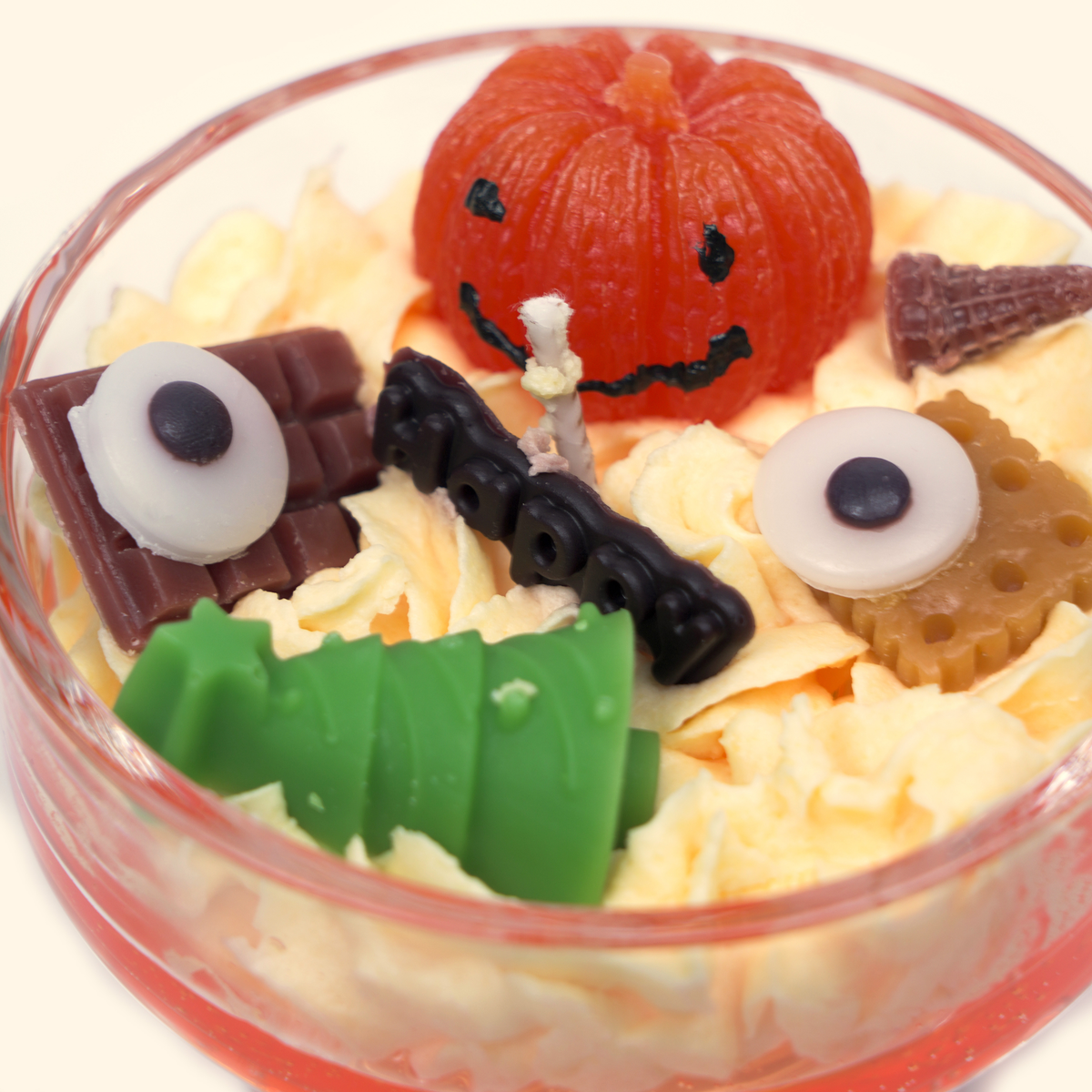 Top close-up of the pumpkin candle with a smiling orange pumpkin, green sculpted wax piece, two eyeball-shaped wax pieces, dark wax detail and cone-shaped decoration.