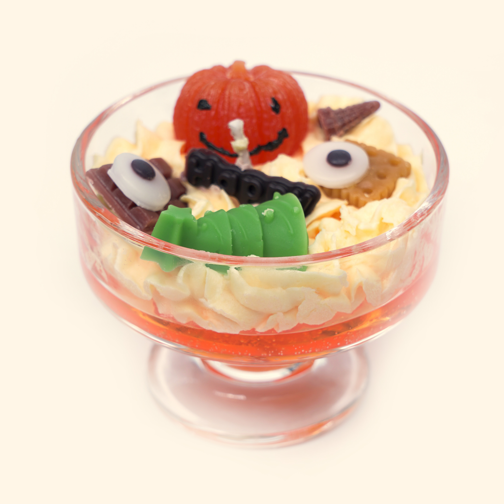 Front view of the Halloween candle with an orange pumpkin centerpiece, green sculpted wax piece, two eyeball-shaped wax pieces and brown candy-inspired details.