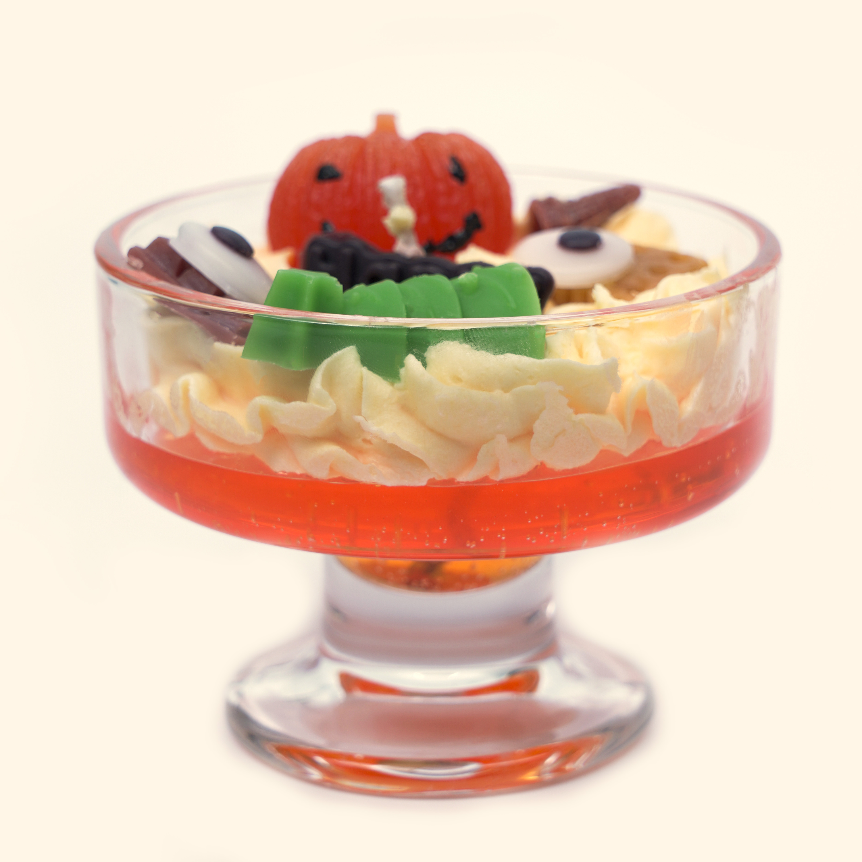 Mischievous Pumpkin Halloween Milkshake Candle in a wide footed glass bowl with a smiling orange pumpkin, green sculpted wax detail, eyeball pieces and cream piped wax over a red gel layer.