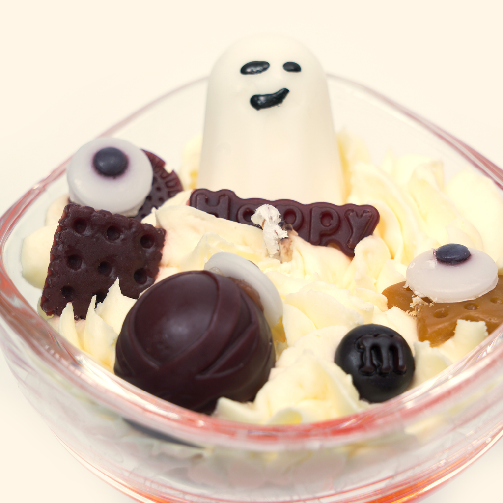 Top close-up of the Ghost Candy House candle with a smiling white ghost, Happy plaque, eyeball-shaped wax pieces, a dark brown round wax piece and biscuit-like wax details.