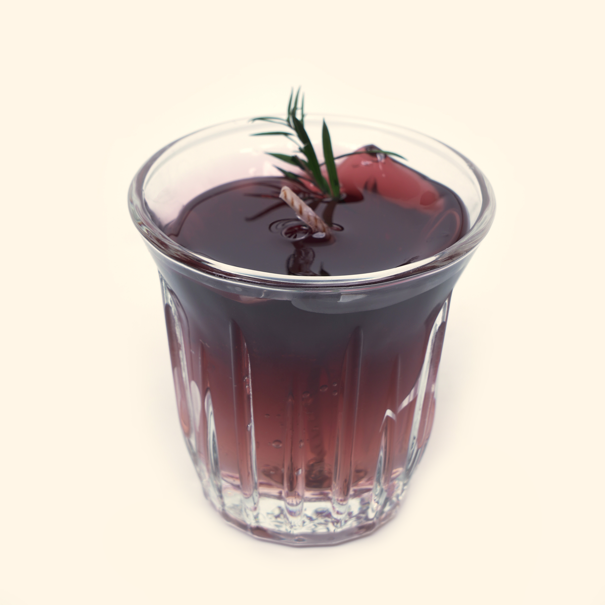 Front view of the coffee candle in a clear vertically fluted glass cup with dark reddish-brown wax and a green sprig.