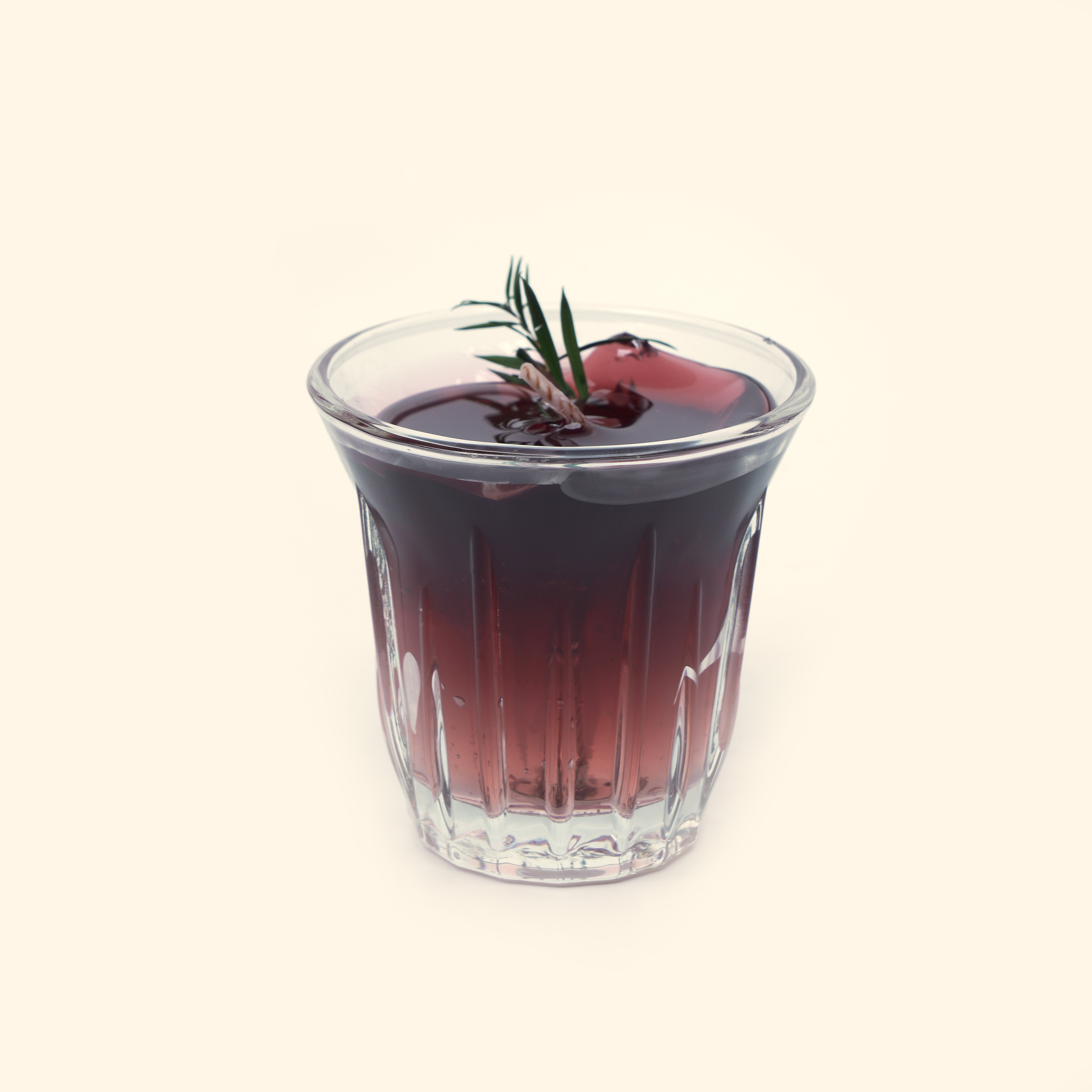 Iced Americano Coffee Candle with dark translucent coffee-colored wax, a twisted wick, green needle-like sprig and ice-cube details in a clear fluted glass cup.