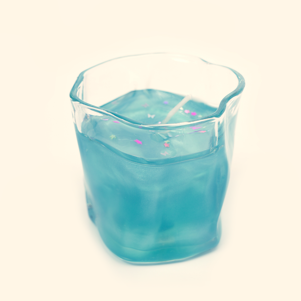 Side view of the blue gel candle in a clear wavy-sided glass cup with colorful star and butterfly confetti suspended near the surface.