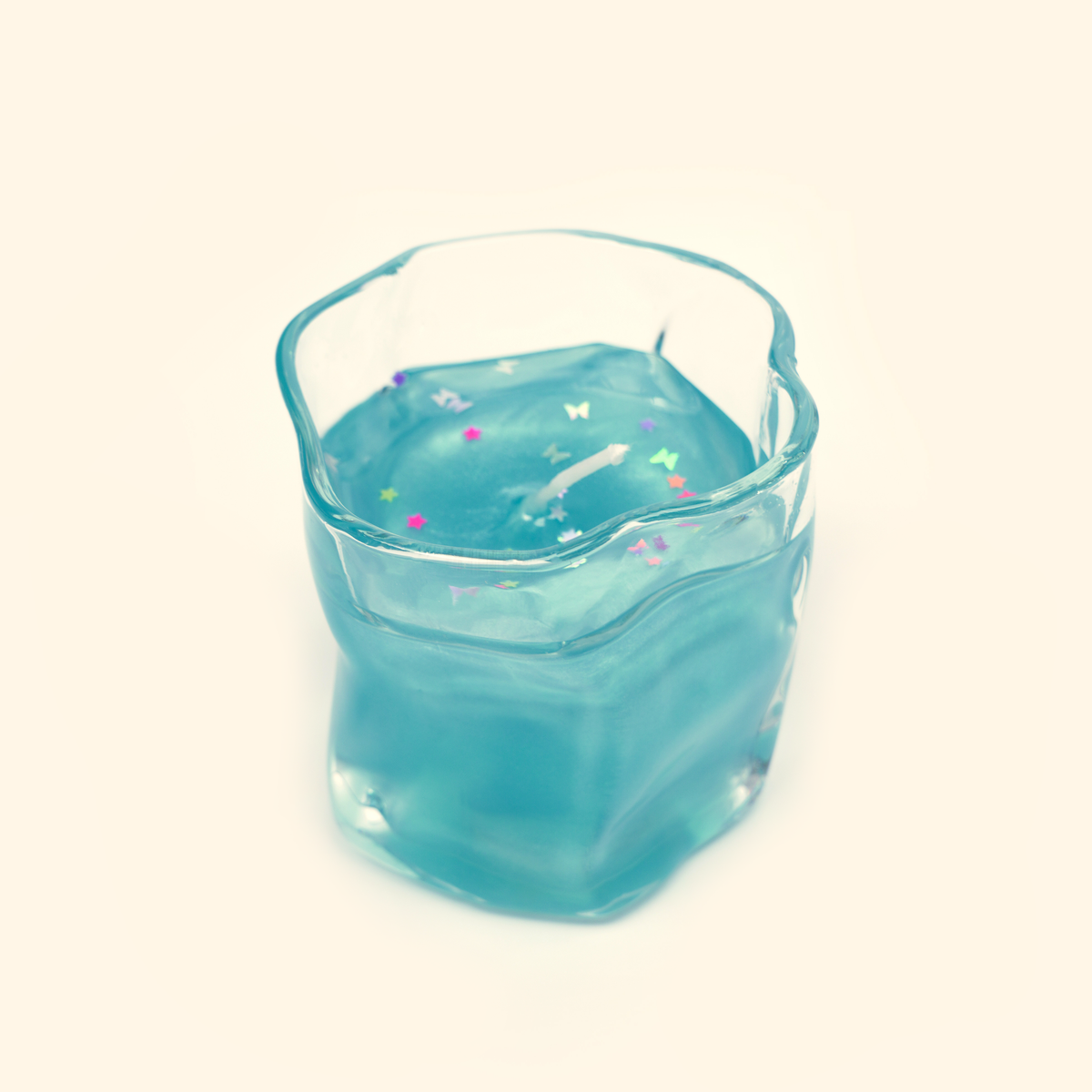 Ocean Dream Gel Candle with translucent aqua-blue gel wax, a white wick and multicolored star and butterfly confetti in a clear wavy glass cup.