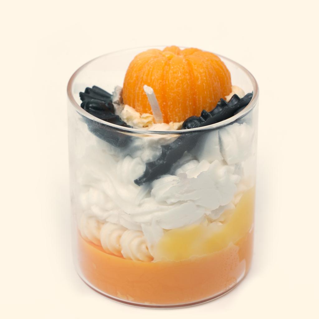 Angled close-up of the pumpkin candle showing black bat wax details, a white wick, cream-colored piped wax and layered orange wax in the glass cup.