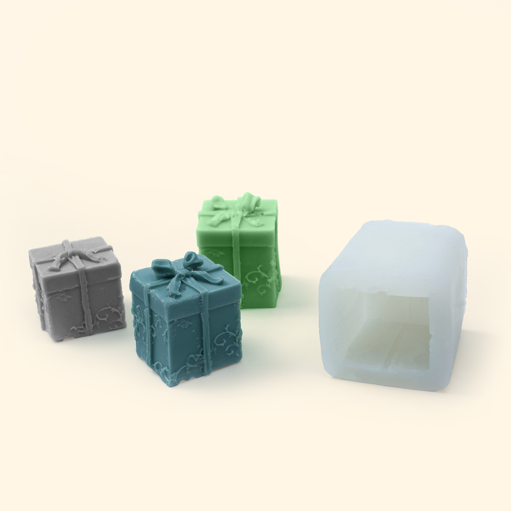 Translucent silicone gift box mold displayed beside sample finished wax pieces showing the sculpted bow and ribbon design.