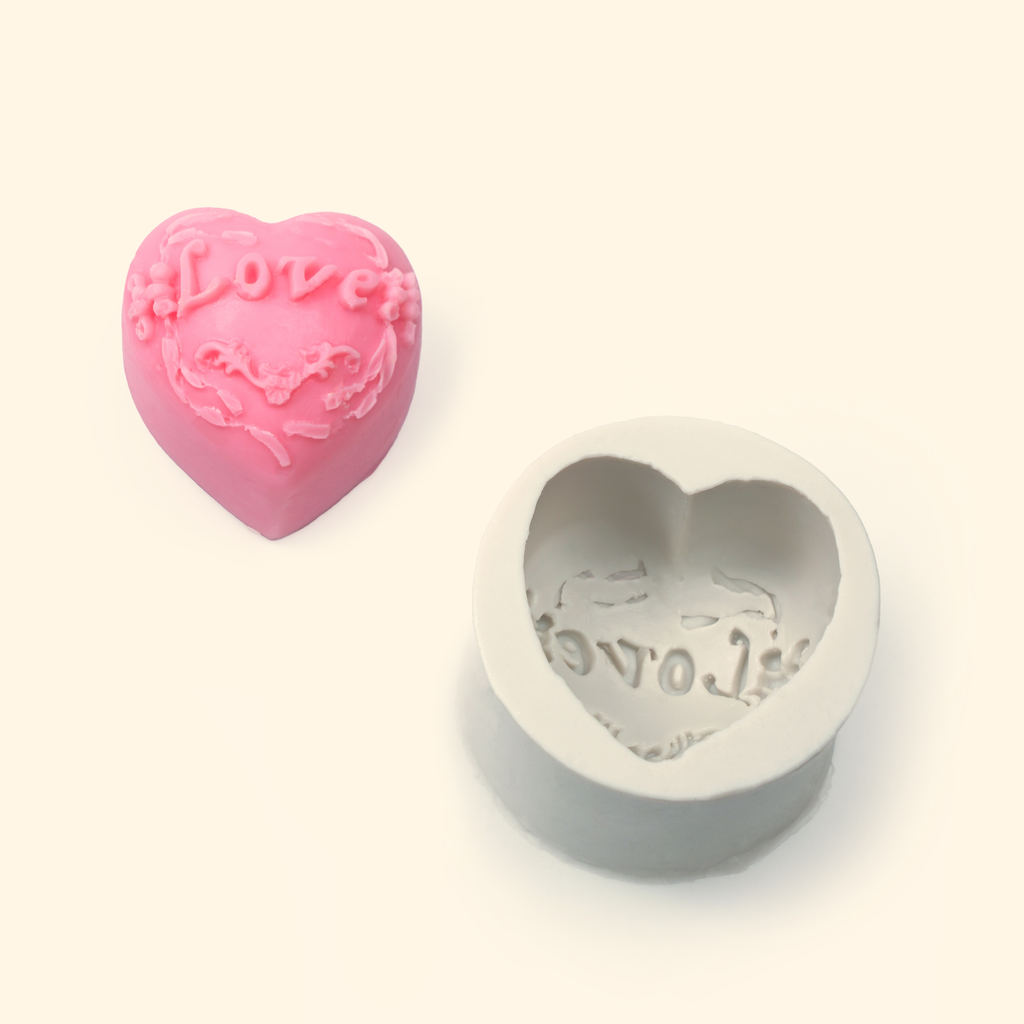 Heart-shaped silicone mold displayed beside a sample finished wax piece showing the embossed LOVE and decorative pattern.