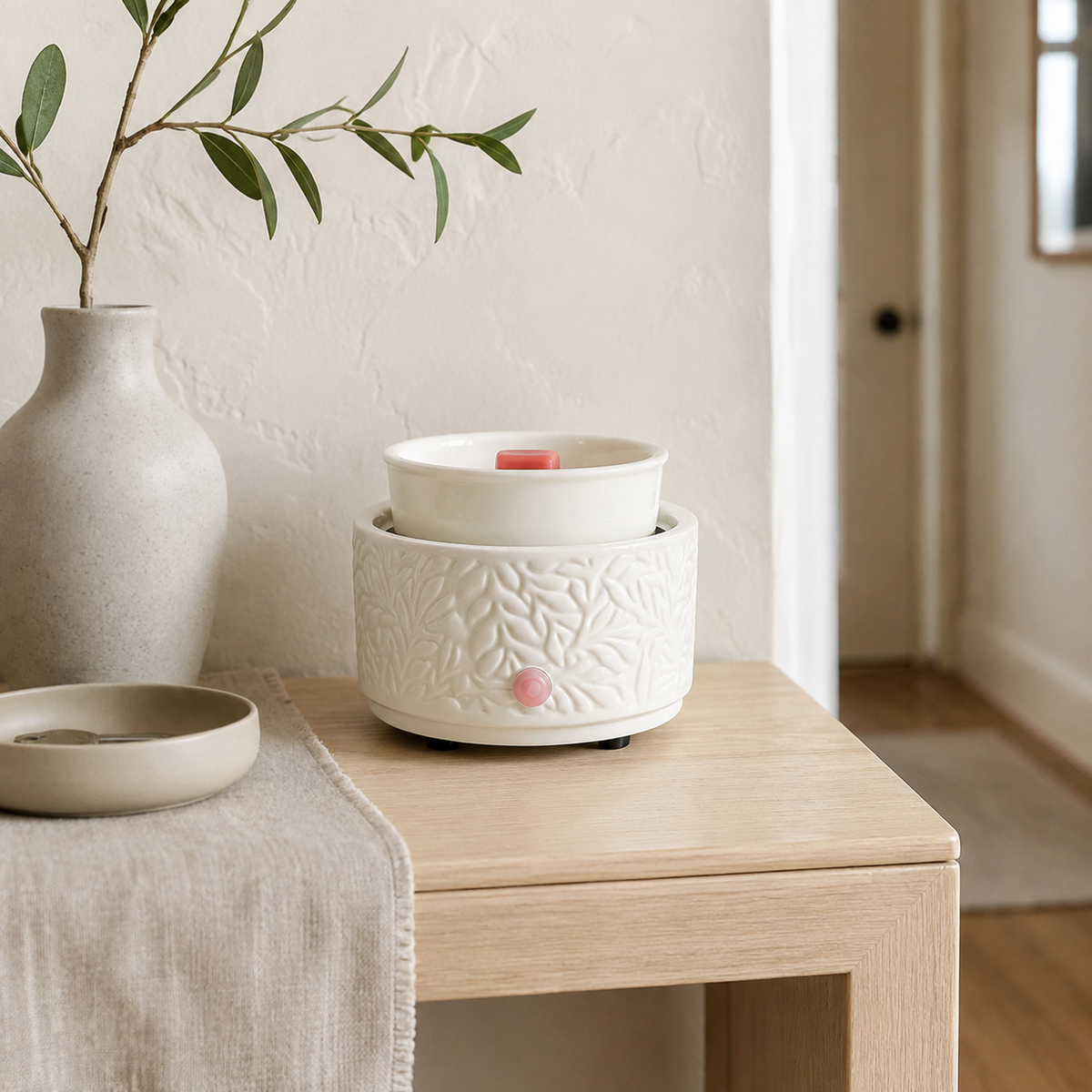 Ivory Botanical tabletop wax warmer with pink wax melt on a light wood console beside a vase and leafy branch.